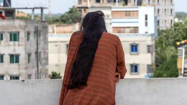 Back shot of a graceful Bengali woman in brown salwar kameez with long black hair, standing on a rooftop. She looks down, a solitary figure in an urban landscape