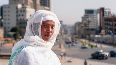 Close-up portrait of an Ethiopian woman wearing a traditional white Habesha kemis with a delicate green-bordered netela, set against the vibrant, sun-drenched backdrop of a modern Addis Ababa street. 
