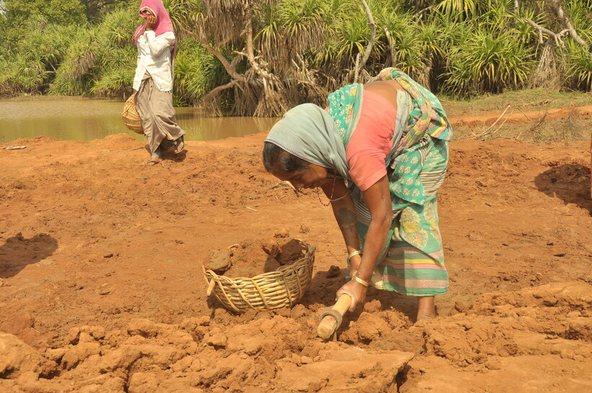 Person digging soil with a hand tool beside a basket in a rural field near a pond, with another person walking in the background
