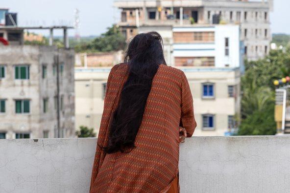 Back shot of a graceful Bengali woman in brown salwar kameez with long black hair, standing on a rooftop. She looks down, a solitary figure in an urban landscape
