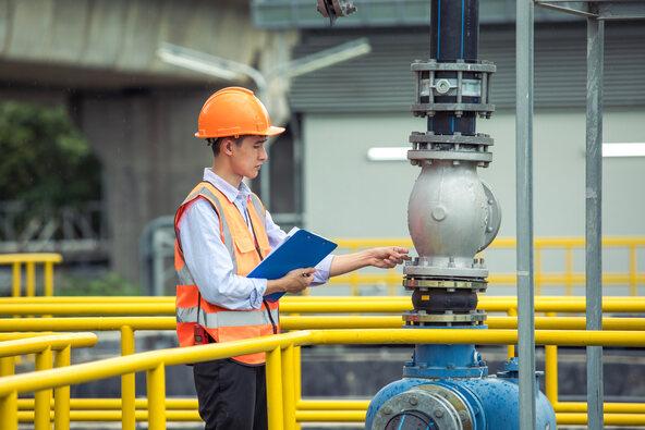 Young Asian male engineer wearing vest and hard hat holding a clipboard inspecting wastewater in an industrial plant