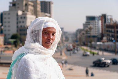 Close-up portrait of an Ethiopian woman wearing a traditional white Habesha kemis with a delicate green-bordered netela, set against the vibrant, sun-drenched backdrop of a modern Addis Ababa street. 