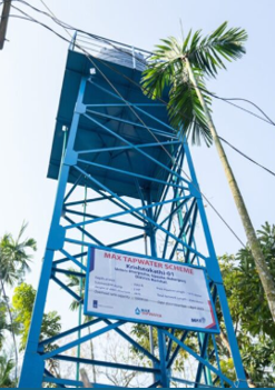 Blue elevated water tank tower with a “MAX TAPWATER SCHEME” sign, surrounded by palm trees
